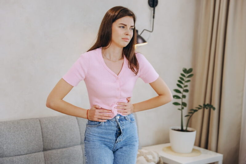 Young woman holding her stomach, struggling with abdominal pain while standing in her living room, reflecting discomfort and distress